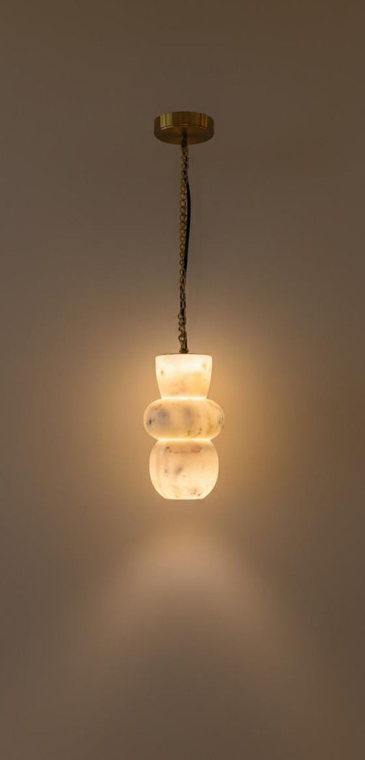 The Tulay Trio is a modern luxury hanging lamp with a gold ceiling mount and chain, showcasing a sculptural frosted white glass shade that softly glows against a beige wall.