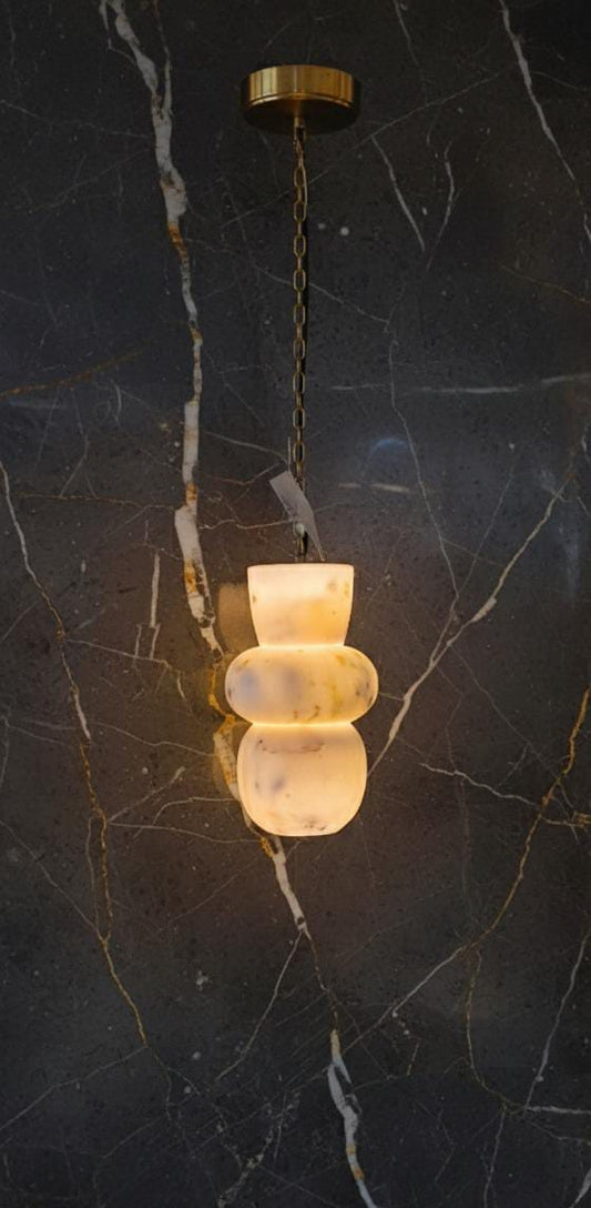 The Tulay Trio is a modern marble pendant light featuring sculptural, frosted white glass shades and a brass chain. Against a dark marble wall with white and gold veining, this luxury lamp glows warmly, adding elegance to any space.