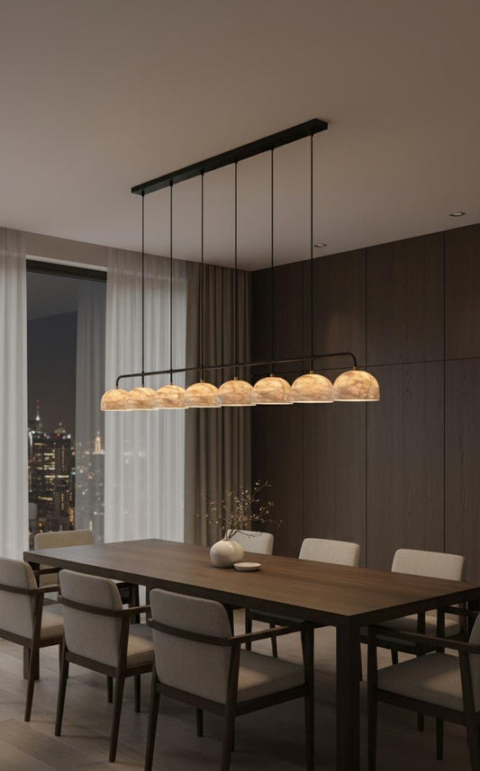 A modern dining room features a long wooden table, eight cushioned chairs, a minimalist vase centerpiece, and the Nova Suspension Chandelier above. Floor-to-ceiling windows showcase a stunning cityscape at night.