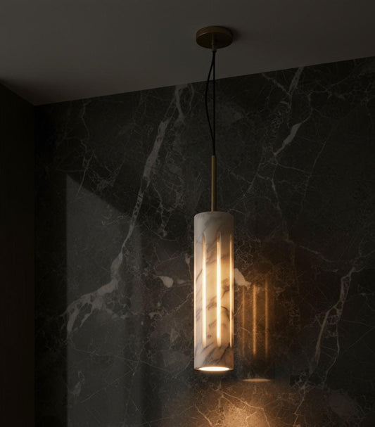 The Cylo cylindrical marble pendant light hangs from the ceiling, casting a warm glow and soft shadows onto a dark marble-patterned wall—an eye-catching sculptural décor piece.