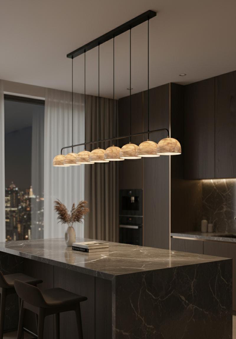 A modern kitchen with dark cabinets, marble countertops, and the Nova Suspension Chandelier with warm globe lamps above the island. A vase with dried plants and stacked books sit on the island, city lights shining through a window.