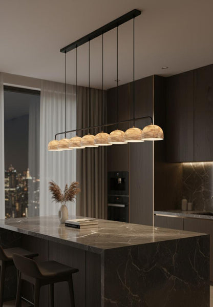 A modern kitchen with dark cabinets, marble countertops, and the Nova Suspension Chandelier with warm globe lamps above the island. A vase with dried plants and stacked books sit on the island, city lights shining through a window.