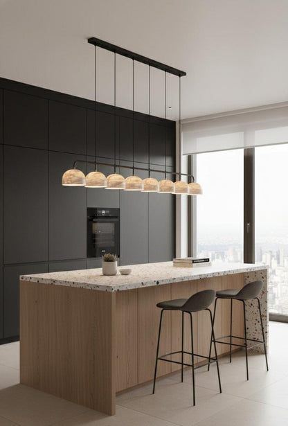 Modern kitchen with matte black cabinets, light wood island, terrazzo countertop, two black bar stools, and modern luxury decor. The Nova Suspension Chandelier highlights the space while a large window frames a city view in the background.